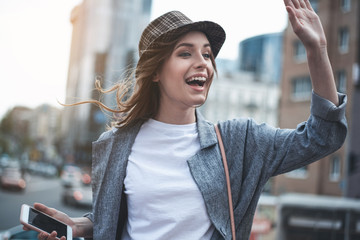 Attractive smiling lady is raising arm to say hello. She is wearing trendy hat and holding...