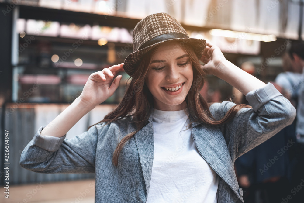 Wall mural cheerful young woman is relaxing outdoors. she is smiling widely and touching trendy hat with both h