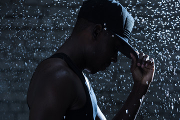 Rapper black man in cap under raindrops at night - obrazy, fototapety, plakaty