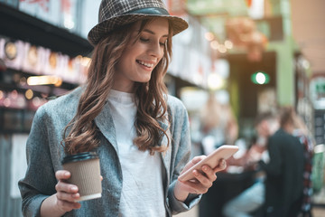 Grinning pretty woman looking at phone. She is standing in trendy hat with cup of hot drink outside