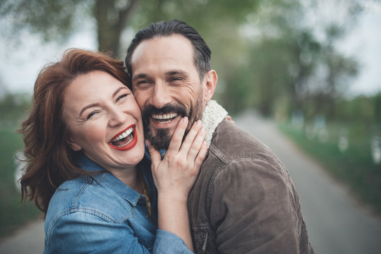 We Are Happy Together. Joyful Husband And Wife Are Bonding To Each Other With Fondness. They Are Looking At Camera And Laughing While Standing On Path Outdoor 