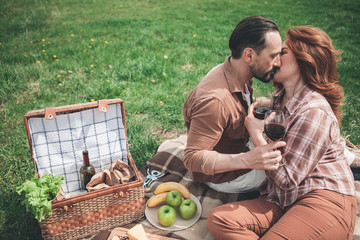 Tender man and woman are kissing in lips with love. Their eyes are closed with fondness. Lovers are sitting on blanket in the nature and holding glasses of wine 