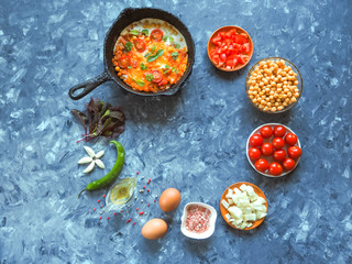 Scrambled eggs with chickpeas and tomatoes on an old pan and a set of vegetables on a grey background.