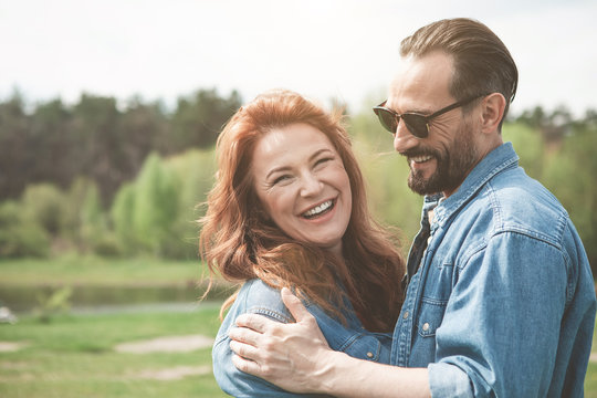 Portrait of joyful red-haired woman enjoying hug of her man and smiling. Picturesque lake is on background. Copy space 