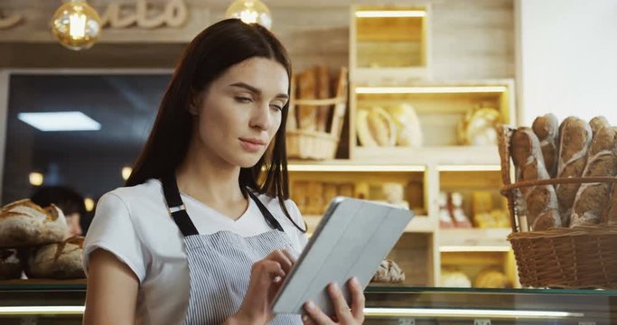 Pretty female baker in glasses scrolling and taping on the tablet computer while standing at the counter in the shop. Indoor