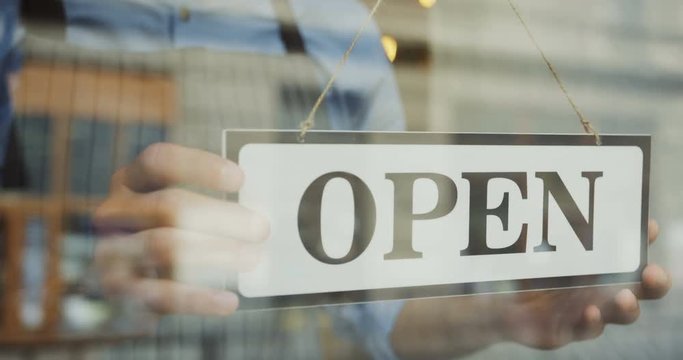 Close up of the male Caucasian hands turning a signboard on the glass door of the shop from CLOSED to OPEN.