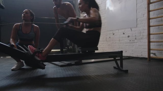 Sportswoman exercising on row machine in gym with trainers. Fit young woman exercising using rowing machine at health club.
