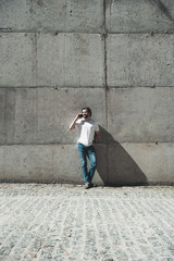 Obraz na płótnie Canvas Pleasant communication. Full length portrait of attractive man in jeans and t shirt standing near concrete wall outside and talking by cellphone. He is smiling
