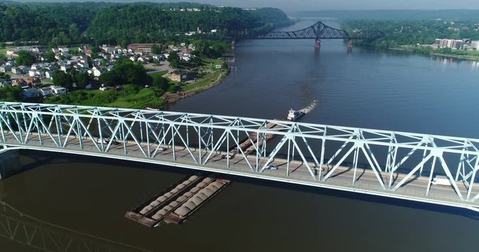 An early morning high angle flyover of a barge traveling on the Ohio River under a bridge. Lowering shot, Pittsburgh suburbs.  	