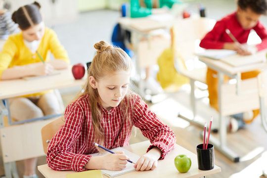 Diligent schoolgirl leaning over her copybook and carrying out individual task at lesson among her classmates
