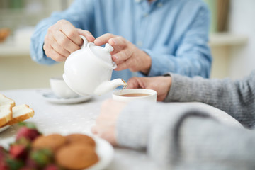 Hands of senior man with white china teapot pouring tea into cup of his wife