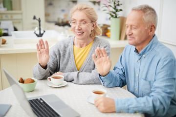 Happy grandparents sitting by table in the kitchen and waving hands to their friends or relatives while talking through video-chat