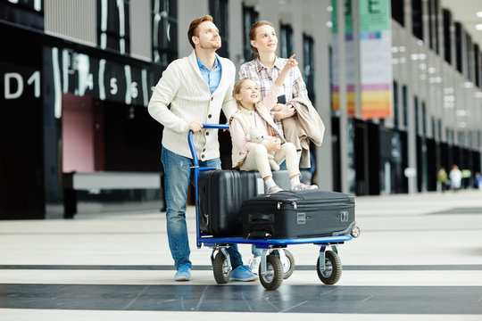 Cute daughter sitting on suitcases on luggage cart pushed by her parents and pointing straight