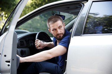 A bearded young man sits in a car with an open door and looks back.