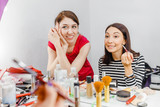 Two young women friends smiling and applying make-up cosmetic at the mirror. Frienship and visage concept
