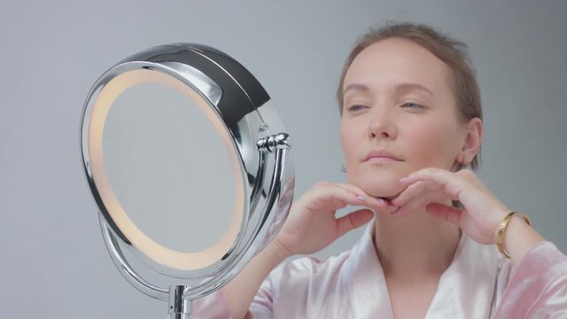 caucasian woman with mirror with ring light with no makeup look examine her skin and makes facial treatment and touching her neck