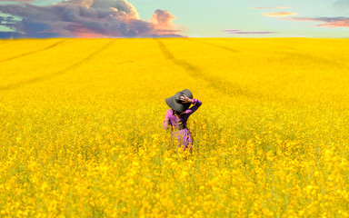 Pretty young woman in violet dress and black hat on blooming sunset rapeseed field