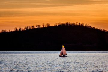 Sailboat on the lake at sunset. Sailing in the evening with beautiful orange light and mountain and forest in the background. The golden hour over the water with a little boat sailing alone.