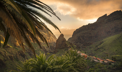Fototapeta na wymiar Masca - Teneriffa - Zentral emporragender Felsen, der umgeben ist von Bergen, Palmen, wenigen Häusern und Grün bei untergehender Sonne mit Wolken 