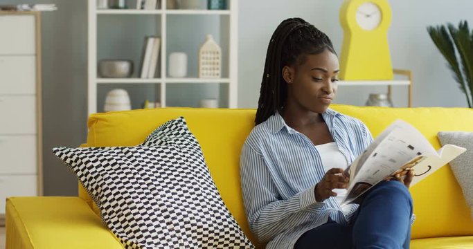 African american woman in the casual clothes flipping pages of the magazine while reading it on the couch un the modern living room. Inside