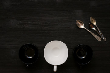 Black and white tea or coffee empty cup and set of silver spoons on black wooden table background with copy space.