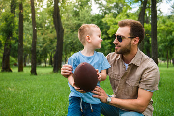 happy father and son looking at each other and holding american football ball at park