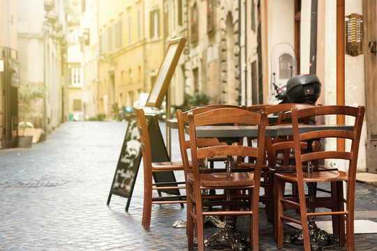 Empty Dining Table With Chairs Is Standing In A Empty Street Lit By Morning Sunlight. Rome, Italy. All Potential Trademarks Are Removed Or Blurred. 