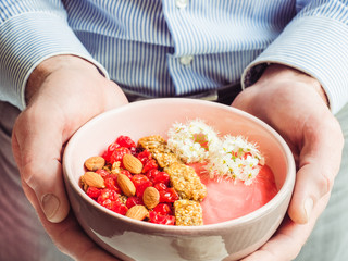 Berry smoothie, muesli with nuts, dry berries, pieces of banana, beautiful, blooming flowers in a pink plate and men's hands. Concept of healthy and tasty food