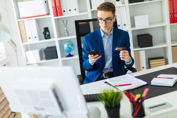 A young man sits at a table in the office and holds a bank card and phone.