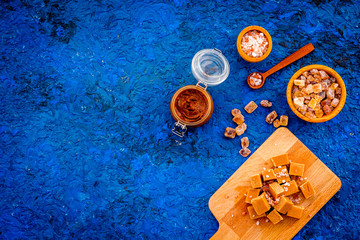 Salted caramel background. Melted caramel in glass jar, caramel cubes on cutting board, sugar and salt on blue desk top view copy space