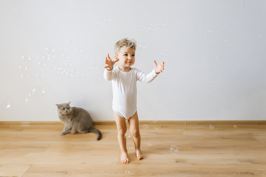 Little Toddler Boy In White Bodysuit Catching Soap Bubbles With Grey Cat Near By At Home