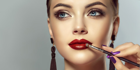  Makeup artist applies  red lipstick  . Beautiful woman face. Hand of make-up master, painting lips...