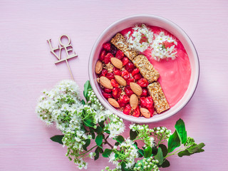Word LOVE, berry smoothie, muesli with nuts, dry berries, pieces of banana in a pink plate, beautiful, blooming flowers, notebook with an inscription on a pink background. Healthy and delicious food