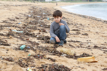 niño triste en la playa mirando la arena llena de plásticos
