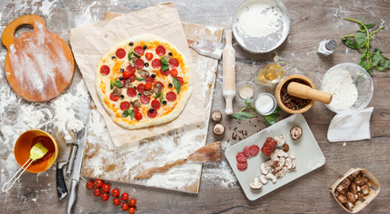 Top view cooking pizza with ingredients, tomatoes, salami and mushrooms on wooden tabletop