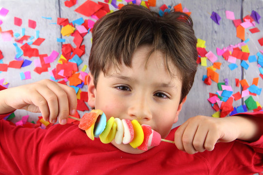 ni&ntilde;o feliz tumbado en el suelo lleno de confeti comiendo una brocheta de coloridas gominolas 