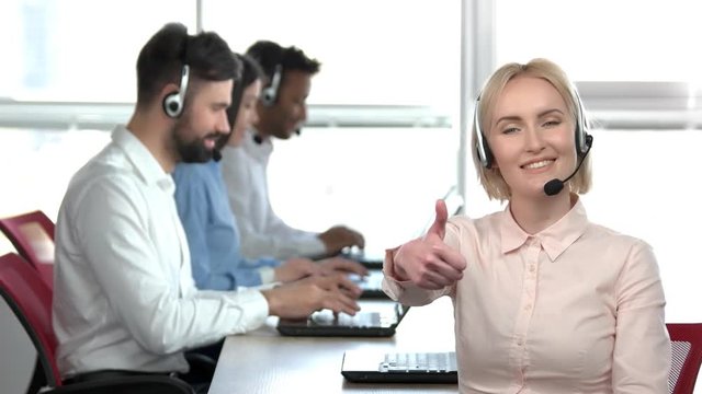 Cheerful blond lady in office showing thumb up. Woman in call center office showing her approval.
