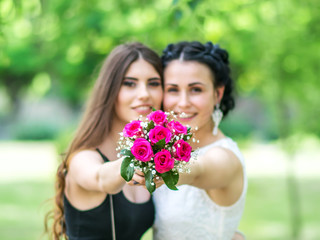Defocused portrait of two beautiful young women holding together wedding bouquet and looking into camera at green park. Bride and bridesmaid holding bouquet on outstretched arms. Focus on the flowers.