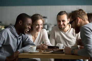 Millennial girl showing funny photos or videos on cellphone to multiracial young friends at cafe meeting, diverse happy friends laughing using mobile phone app together sitting at coffee house table