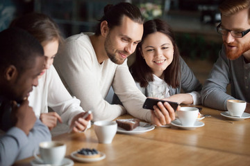 Diverse happy friends having fun using smartphone apps online hanging together in cafe, multiracial...