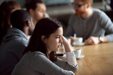 Frustrated upset millennial girl sitting alone at cafe table after conflict ignoring friends, young...