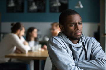 Frustrated excluded outstand african american man suffers from bullying or racial discrimination having no friends sitting alone in cafe, sad depressed black guy upset being rejected by white people - obrazy, fototapety, plakaty