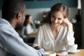 Happy interracial couple flirting talking sitting at cafe table, african man holding hand of...