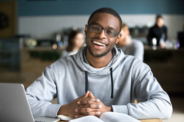 Obraz premium Happy african casual man in glasses sits at cafe table looking at camera, cheerful black student posing studying with book in coffee house, smiling businessman works on laptop in cafeteria, portrait