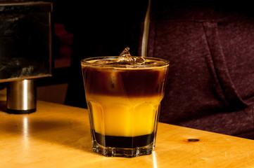 Preparation of a colorful cocktail of bumble or crazy bee with orange juice caramel syrup and espresso coffee in a glass cup on a wooden table coffee maker on the background