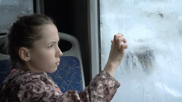 A little girl sits in the bus in the back seat and draws pictures with her finger on the wet misted glass. Outside the window drive cars. Portrait. Close up. High detail. 4K, 25 fps