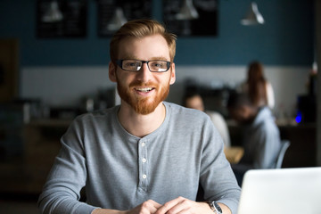 Smiling redhead man with laptop looking at camera in cafe, happy millennial guy in glasses posing in cafeteria working or studying in coffee shop, young businessman entrepreneur freelancer portrait
