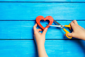 Female hands cut a wooden red heart with scissors on a blue background. The concept of breaking relations, quarrel, divorce. The pair diverges. Betrayal of the other, the patient separation.