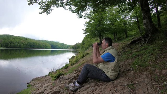 Man looks through binoculars at distance while sitting at scenic river bank on green grass in shadow of tree.