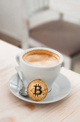 Cryptocurrency golden bitcoin standing on coffee cup. Golden cryptocurrency and white cup hot coffee mug on the soft background.